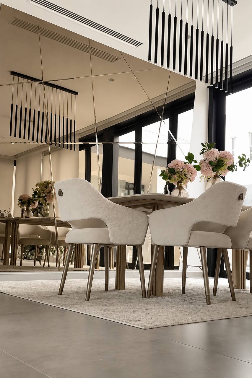Comedor con muro espejado en tono bronce, sillas beige tapizadas y lámpara colgante de tubos negros